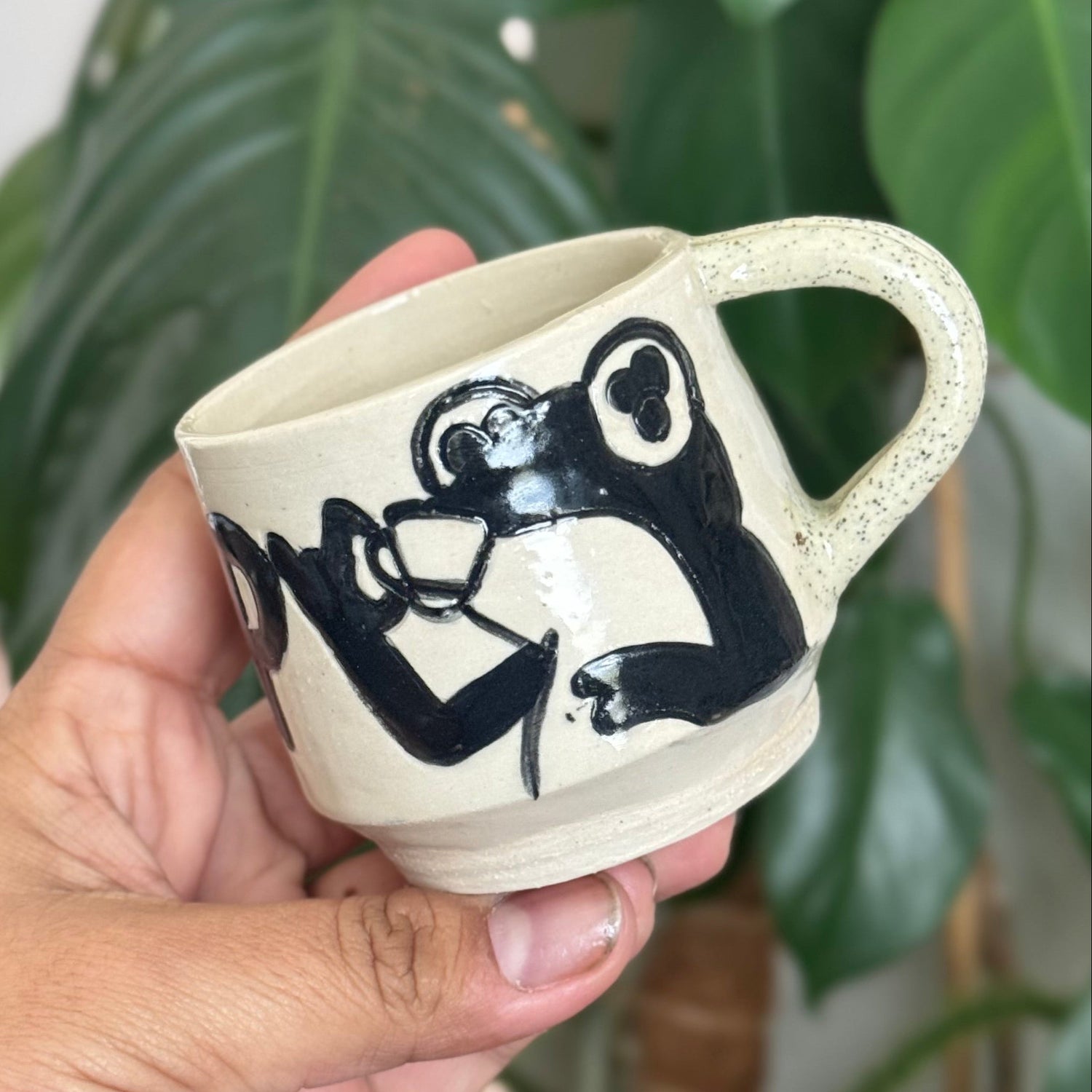 Hand holding a ceramic mug with a black and white design against a green plant background