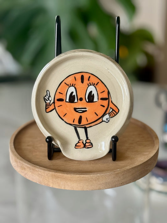Miss Minutes spoon rest front view showing hand-illustrated orange cartoon clock character on cream speckled glaze
